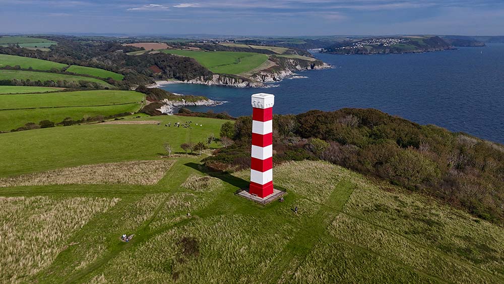 A tall red and white striped lighthouse stands on a green hillside, overlooking the coastline and ocean under a clear blue sky.