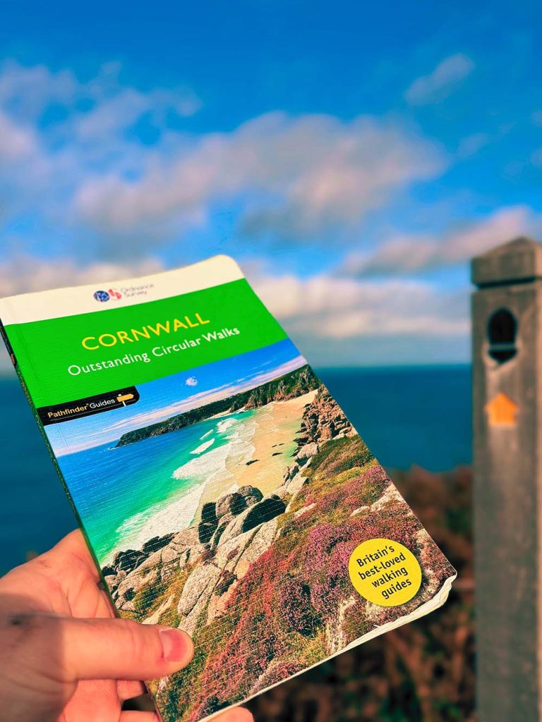 A person holding a guidebook titled 'Cornwall: Outstanding Circular Walks' against a scenic backdrop of the ocean and sky.