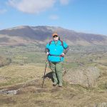 A person standing on a rocky outcrop with a scenic view of rolling hills and mountains in the background under a clear blue sky.