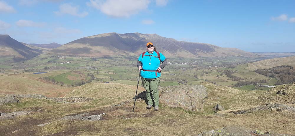 A person standing on a rocky outcrop with a scenic view of rolling hills and mountains in the background under a clear blue sky.
