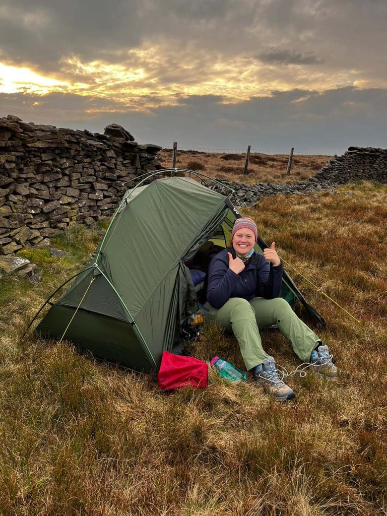 A woman sits outside a green camping tent, giving a thumbs-up in a grassy area with a stone wall in the background. The sky is cloudy, indicating a sunset atmosphere.