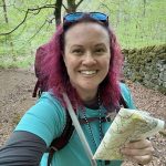 Rebecca Dawson with curly pink hair wearing a turquoise shirt, holding a map and smiling, in a green forest setting.