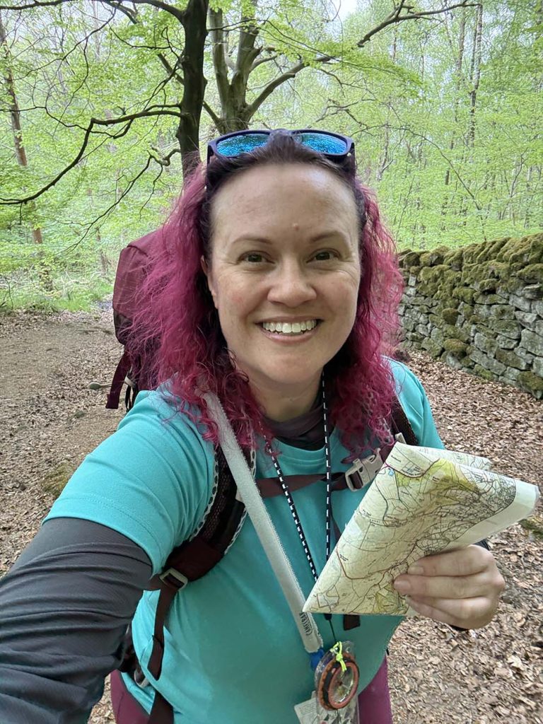 Rebecca Dawson with curly pink hair wearing a turquoise shirt, holding a map and smiling, in a green forest setting.