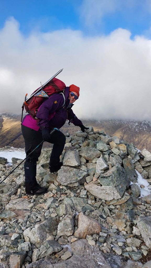 A person in outdoor gear, including a purple jacket and red beanie, climbs a rocky peak, looking back at the camera with a smile. The landscape is mountainous with clouds in the background.
